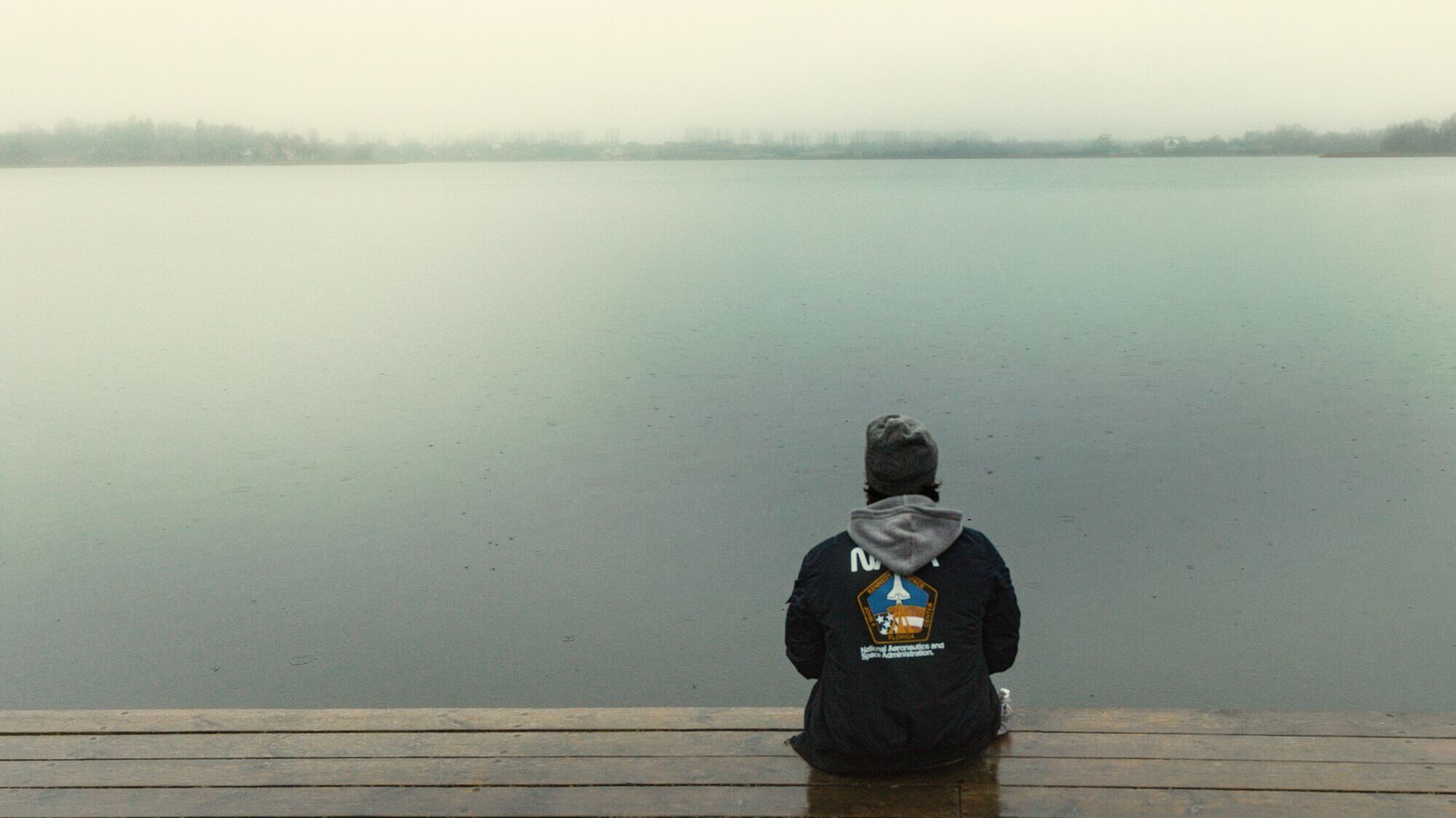 A man in a beanie and hoodie sitting at the end of a dock, facing a huge, calm, misty lake